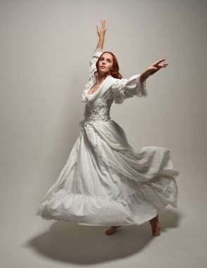 Tam boy, kızıl saçlı, tarihi fantezi gelinlik giyen bir model. Peri masalı balosu elbisesi. Beyaz stüdyo arka planında akan kumaşla izole edilmiş dans figürü