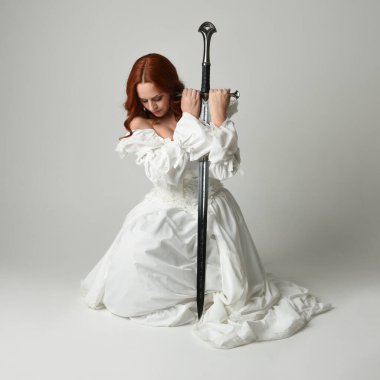 Tam boy portre, tarihi beyaz fantezi gelinlik giyen güzel kadın manken, kılıç tutan peri masalı balosu elbisesi. Stüdyonun arka planında izole bir poz.