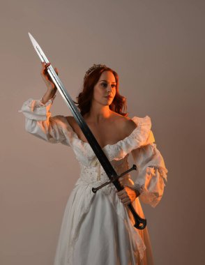 Güzel, kızıl saçlı, tarihi, beyaz, fantezi bir gelinlik, peri masalı balosu elbisesi ve kılıç tutan altın taç giymiş güzel bir manken. Stüdyo arkaplanındaki izole kişi.