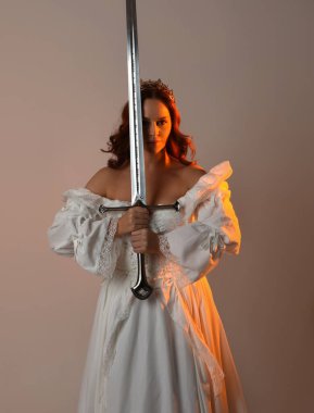 Güzel, kızıl saçlı, tarihi, beyaz, fantezi bir gelinlik, peri masalı balosu elbisesi ve kılıç tutan altın taç giymiş güzel bir manken. Stüdyo arkaplanındaki izole kişi.