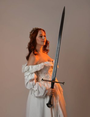 Güzel, kızıl saçlı, tarihi, beyaz, fantezi bir gelinlik, peri masalı balosu elbisesi ve kılıç tutan altın taç giymiş güzel bir manken. Stüdyo arkaplanındaki izole kişi.