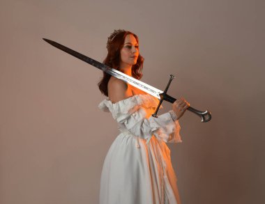 Güzel, kızıl saçlı, tarihi, beyaz, fantezi bir gelinlik, peri masalı balosu elbisesi ve kılıç tutan altın taç giymiş güzel bir manken. Stüdyo arkaplanındaki izole kişi.