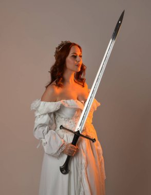 Güzel, kızıl saçlı, tarihi, beyaz, fantezi bir gelinlik, peri masalı balosu elbisesi ve kılıç tutan altın taç giymiş güzel bir manken. Stüdyo arkaplanındaki izole kişi.