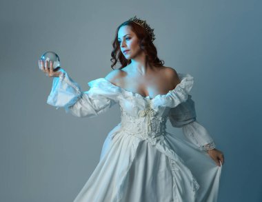 Yakın plan, güzel kızıl saçlı, tarihi beyaz fantezi gelinlik giyen, peri masalı elbisesi giyen, kristal kahin küresi tutan izole bir figür. Stüdyo arka planında mavi karamsar ışıklandırma