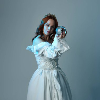 Yakın plan, güzel kızıl saçlı, tarihi beyaz fantezi gelinlik giyen, peri masalı elbisesi giyen, kristal kahin küresi tutan izole bir figür. Stüdyo arka planında mavi karamsar ışıklandırma