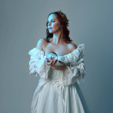 Yakın plan, güzel kızıl saçlı, tarihi beyaz fantezi gelinlik giyen, peri masalı elbisesi giyen, kristal kahin küresi tutan izole bir figür. Stüdyo arka planında mavi karamsar ışıklandırma