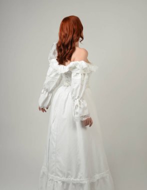 Yakın plan güzel kızıl saçlı manken tarihi fantezi gelinlik giyiyor, peri masalı balosu elbisesi giyiyor. Kameradan uzaklaşmak, geriye bakmak, beyaz stüdyo arka planında soyutlanmış bir figür.