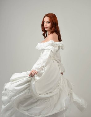 Yakın plan güzel kızıl saçlı manken tarihi fantezi gelinlik giyiyor, peri masalı balosu elbisesi giyiyor. Kameradan uzaklaşmak, geriye bakmak, beyaz stüdyo arka planında soyutlanmış bir figür.