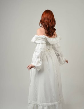 Yakın plan güzel kızıl saçlı manken tarihi fantezi gelinlik giyiyor, peri masalı balosu elbisesi giyiyor. Kameradan uzaklaşmak, geriye bakmak, beyaz stüdyo arka planında soyutlanmış bir figür.