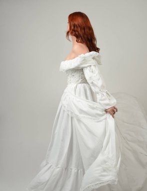 Yakın plan güzel kızıl saçlı manken tarihi fantezi gelinlik giyiyor, peri masalı balosu elbisesi giyiyor. Kameradan uzaklaşmak, geriye bakmak, beyaz stüdyo arka planında soyutlanmış bir figür.