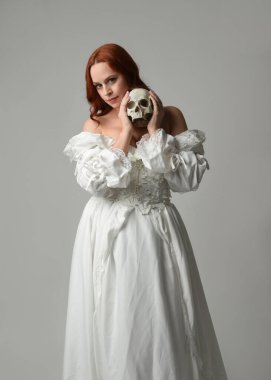 Güzel, kızıl saçlı, tarihi, beyaz, fantezi bir gelinlik giyen, peri masalı balosu elbisesi giyen iskelet kafa tutan güzel bir manken. Beyaz stüdyo arka planında izole bir figür.