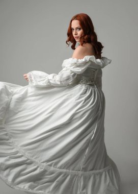 Yakın plan güzel kızıl saçlı manken tarihi fantezi gelinlik giyiyor, peri masalı balosu elbisesi giyiyor. Kameradan uzaklaşmak, geriye bakmak, beyaz stüdyo arka planında soyutlanmış bir figür.