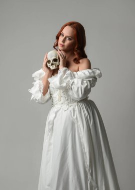 Güzel, kızıl saçlı, tarihi, beyaz, fantezi bir gelinlik giyen, peri masalı balosu elbisesi giyen iskelet kafa tutan güzel bir manken. Beyaz stüdyo arka planında izole bir figür.