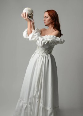 Güzel, kızıl saçlı, tarihi, beyaz, fantezi bir gelinlik giyen, peri masalı balosu elbisesi giyen iskelet kafa tutan güzel bir manken. Beyaz stüdyo arka planında izole bir figür.