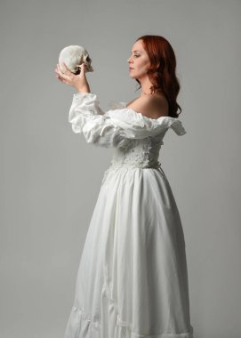 Güzel, kızıl saçlı, tarihi, beyaz, fantezi bir gelinlik giyen, peri masalı balosu elbisesi giyen iskelet kafa tutan güzel bir manken. Beyaz stüdyo arka planında izole bir figür.