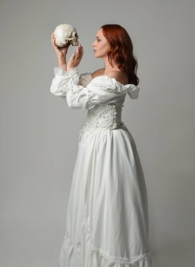 Güzel, kızıl saçlı, tarihi, beyaz, fantezi bir gelinlik giyen, peri masalı balosu elbisesi giyen iskelet kafa tutan güzel bir manken. Beyaz stüdyo arka planında izole bir figür.