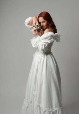 Güzel, kızıl saçlı, tarihi, beyaz, fantezi bir gelinlik giyen, peri masalı balosu elbisesi giyen iskelet kafa tutan güzel bir manken. Beyaz stüdyo arka planında izole bir figür.