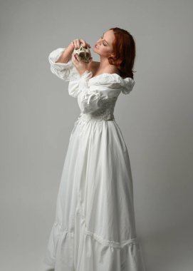 Güzel, kızıl saçlı, tarihi, beyaz, fantezi bir gelinlik giyen, peri masalı balosu elbisesi giyen iskelet kafa tutan güzel bir manken. Beyaz stüdyo arka planında izole bir figür.