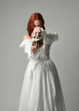 Güzel, kızıl saçlı, tarihi, beyaz, fantezi bir gelinlik giyen, peri masalı balosu elbisesi giyen iskelet kafa tutan güzel bir manken. Beyaz stüdyo arka planında izole bir figür.