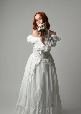 Güzel, kızıl saçlı, tarihi, beyaz, fantezi bir gelinlik giyen, peri masalı balosu elbisesi giyen iskelet kafa tutan güzel bir manken. Beyaz stüdyo arka planında izole bir figür.