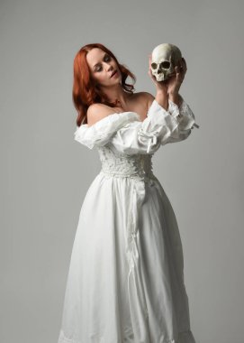 Güzel, kızıl saçlı, tarihi, beyaz, fantezi bir gelinlik giyen, peri masalı balosu elbisesi giyen iskelet kafa tutan güzel bir manken. Beyaz stüdyo arka planında izole bir figür.