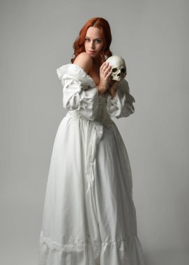 Güzel, kızıl saçlı, tarihi, beyaz, fantezi bir gelinlik giyen, peri masalı balosu elbisesi giyen iskelet kafa tutan güzel bir manken. Beyaz stüdyo arka planında izole bir figür.