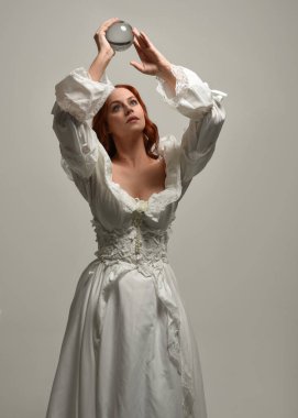 Yakın plan güzel kızıl saçlı manken tarihi beyaz fantezi gelinlik giyiyor, peri masalı elbisesi giyiyor, elinde kristal kahinler var. Beyaz arkaplanda yukarıdaki stüdyonun ışığı