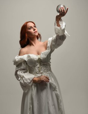 Yakın plan güzel kızıl saçlı manken tarihi beyaz fantezi gelinlik giyiyor, peri masalı elbisesi giyiyor, elinde kristal kahinler var. Beyaz arkaplanda yukarıdaki stüdyonun ışığı