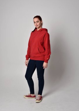 Çağdaş güncel kıyafetler giyen kadın modelin tam boy figürü kırmızı kapüşonlu kazak, dar kot pantolon ve spor ayakkabılar. Stüdyonun arka planında izole bir şekilde kameraya doğru yürüyor..