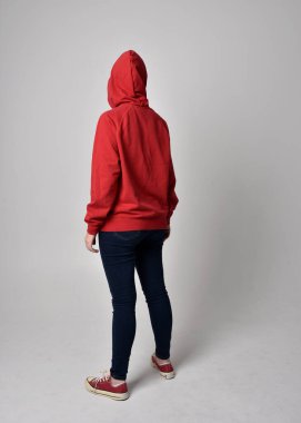 Çağdaş güncel kıyafetler giyen kadın modelin tam boy figürü kırmızı kapüşonlu kazak, dar kot pantolon, spor ayakkabılar. Duruş uzaklaşıyor. Arka plan, izole edilmiş beyaz stüdyo arka plan..