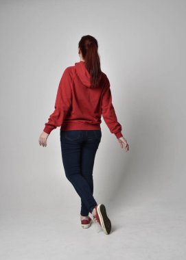 Çağdaş güncel kıyafetler giyen kadın modelin tam boy figürü kırmızı kapüşonlu kazak, dar kot pantolon, spor ayakkabılar. Duruş uzaklaşıyor. Arka plan, izole edilmiş beyaz stüdyo arka plan..