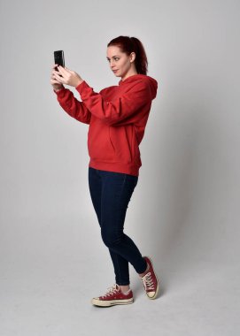 Çağdaş güncel kıyafetler giyen kadın modelin tam boy figürü kırmızı kapüşonlu kazak, dar kot pantolon, spor ayakkabılar. Ayakta duran poz cep telefonu, izole edilmiş beyaz stüdyo arka planı.