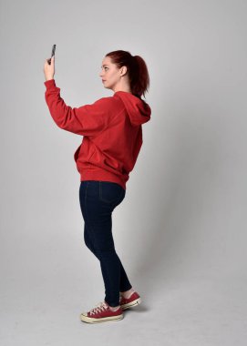Çağdaş güncel kıyafetler giyen kadın modelin tam boy figürü kırmızı kapüşonlu kazak, dar kot pantolon, spor ayakkabılar. Ayakta duran poz cep telefonu, izole edilmiş beyaz stüdyo arka planı.