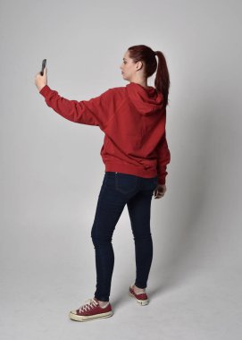 Çağdaş güncel kıyafetler giyen kadın modelin tam boy figürü kırmızı kapüşonlu kazak, dar kot pantolon, spor ayakkabılar. Ayakta duran poz cep telefonu, izole edilmiş beyaz stüdyo arka planı.