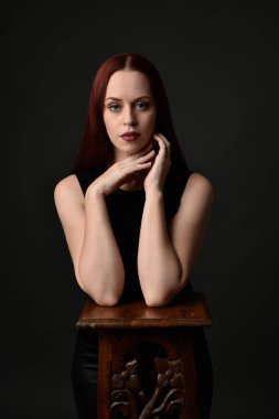 Uzun kızıl saçlı, siyah gömlekli, el ve kol hareketleriyle ahşap çizgilere yaslanmış güzel bir kadın modelin portresi. Koyu arkaplanda klasik stüdyo resmi.