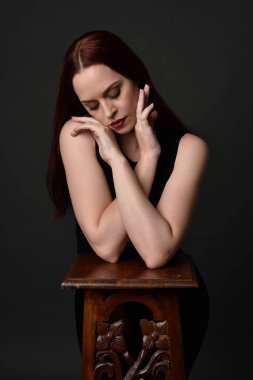 Uzun kızıl saçlı, siyah gömlekli, el ve kol hareketleriyle ahşap çizgilere yaslanmış güzel bir kadın modelin portresi. Koyu arkaplanda klasik stüdyo resmi.