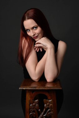Uzun kızıl saçlı, siyah gömlekli, el ve kol hareketleriyle ahşap çizgilere yaslanmış güzel bir kadın modelin portresi. Koyu arkaplanda klasik stüdyo resmi.