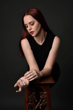 Uzun kızıl saçlı, siyah gömlekli, el ve kol hareketleriyle ahşap çizgilere yaslanmış güzel bir kadın modelin portresi. Koyu arkaplanda klasik stüdyo resmi.