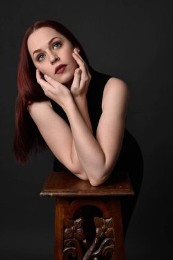 Uzun kızıl saçlı, siyah gömlekli, el ve kol hareketleriyle ahşap çizgilere yaslanmış güzel bir kadın modelin portresi. Koyu arkaplanda klasik stüdyo resmi.