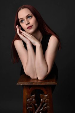 Uzun kızıl saçlı, siyah gömlekli, el ve kol hareketleriyle ahşap çizgilere yaslanmış güzel bir kadın modelin portresi. Koyu arkaplanda klasik stüdyo resmi.