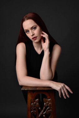 Uzun kızıl saçlı, siyah gömlekli, el ve kol hareketleriyle ahşap çizgilere yaslanmış güzel bir kadın modelin portresi. Koyu arkaplanda klasik stüdyo resmi.