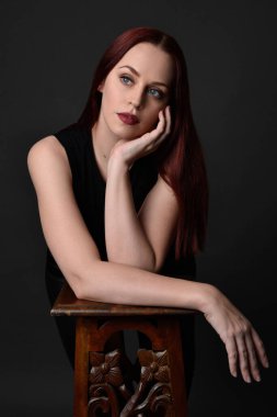 Uzun kızıl saçlı, siyah gömlekli, el ve kol hareketleriyle ahşap çizgilere yaslanmış güzel bir kadın modelin portresi. Koyu arkaplanda klasik stüdyo resmi.