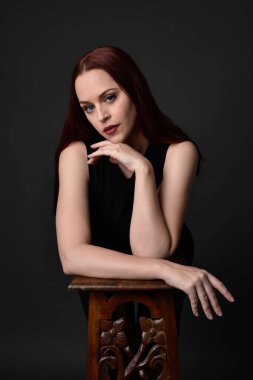 Uzun kızıl saçlı, siyah gömlekli, el ve kol hareketleriyle ahşap çizgilere yaslanmış güzel bir kadın modelin portresi. Koyu arkaplanda klasik stüdyo resmi.