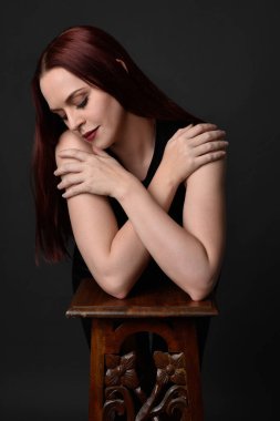 Uzun kızıl saçlı, siyah gömlekli, el ve kol hareketleriyle ahşap çizgilere yaslanmış güzel bir kadın modelin portresi. Koyu arkaplanda klasik stüdyo resmi.