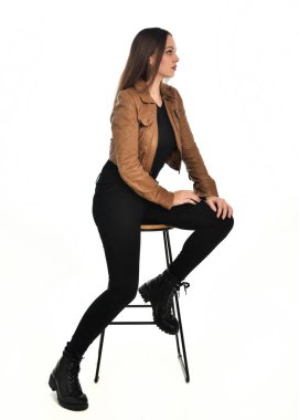 Kahverengi deri ceketli, modern siyah kıyafetler giyen güzel esmer kadın modelin tam boy portresi. Beyaz stüdyo arka planında izole bir figür..