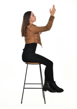 Kahverengi deri ceketli, modern siyah kıyafetler giyen güzel esmer kadın modelin tam boy portresi. Beyaz stüdyo arka planında izole bir figür..