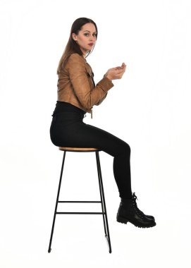 Kahverengi deri ceketli, modern siyah kıyafetler giyen güzel esmer kadın modelin tam boy portresi. Beyaz stüdyo arka planında izole bir figür..