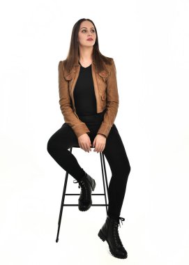 Kahverengi deri ceketli, modern siyah kıyafetler giyen güzel esmer kadın modelin tam boy portresi. Beyaz stüdyo arka planında izole bir figür..