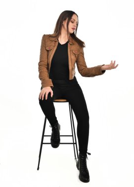Kahverengi deri ceketli, modern siyah kıyafetler giyen güzel esmer kadın modelin tam boy portresi. Beyaz stüdyo arka planında izole bir figür..