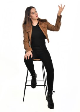 Kahverengi deri ceketli, modern siyah kıyafetler giyen güzel esmer kadın modelin tam boy portresi. Beyaz stüdyo arka planında izole bir figür..